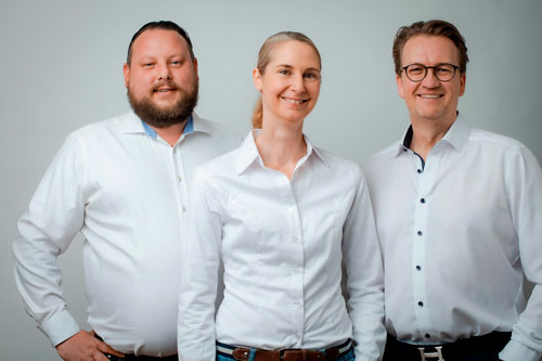 Eine Frau und zwei Männer posieren lächelnd in weißen Hemden