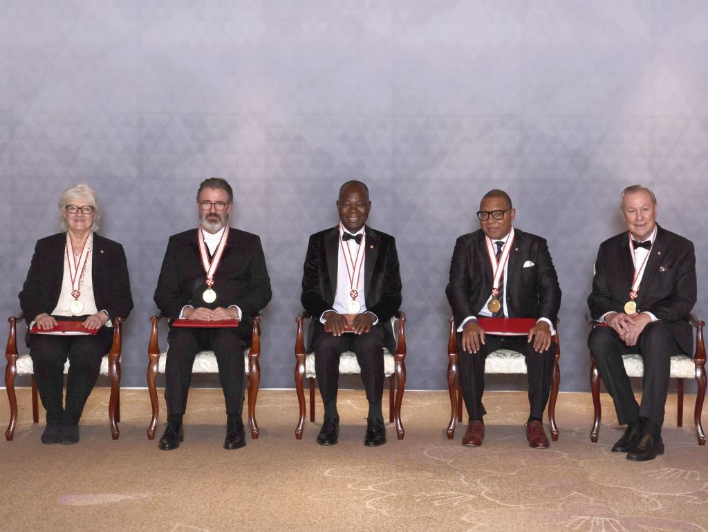 Fünf Männer in Anzügen sitzen auf Stühlen und zeigen stolz ihre Medaillen.