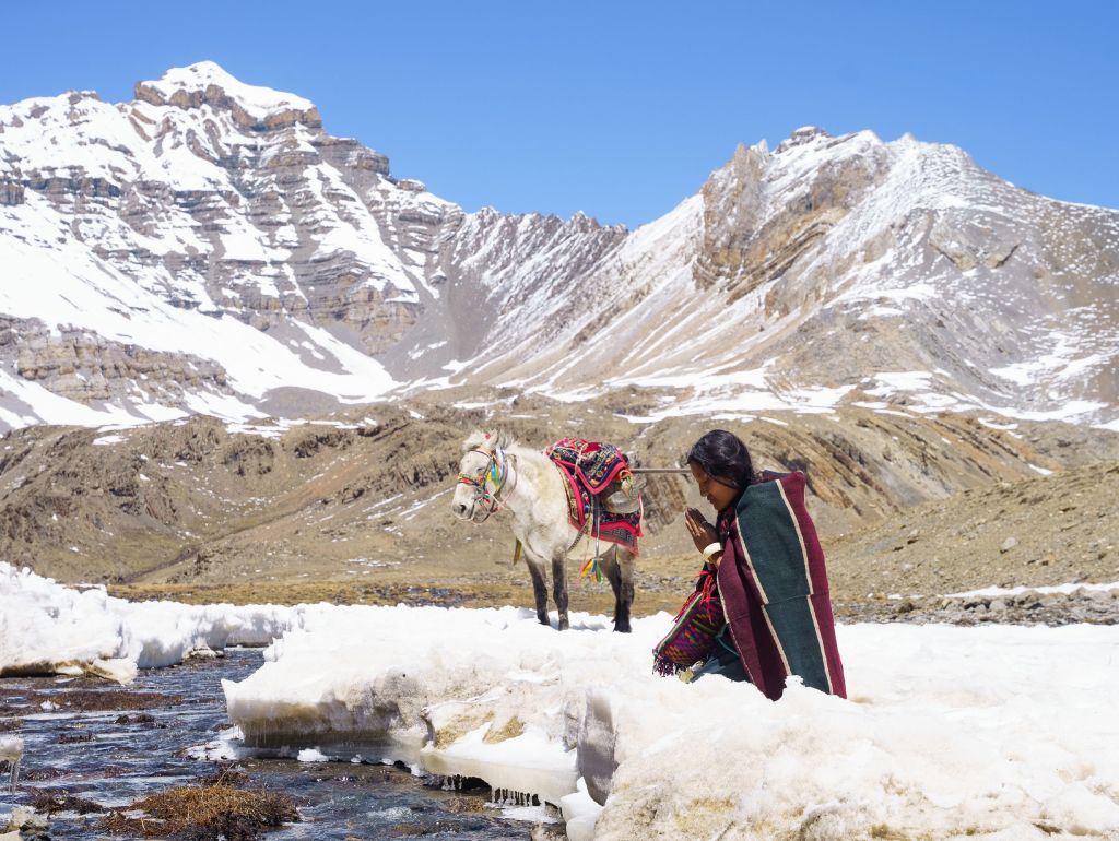 Ein Mensch im Schnee mit seinem Pferd am Ufer eines Baches im Hintergrund hohe Berge