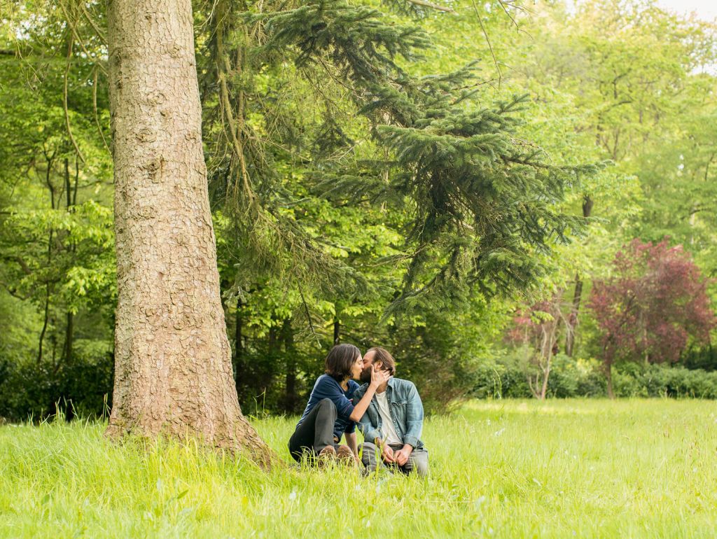 ein Pärchen sitzt küssend auf einer Wiese vor einem Baum 