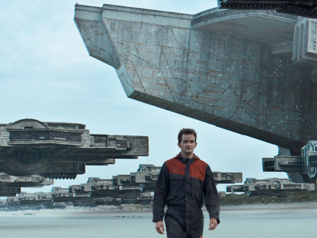 Ein Mann in Futuristischer Kleidung auf einem Flugplatz mit Mehreren Großen Raumschiffen im Hintergrund