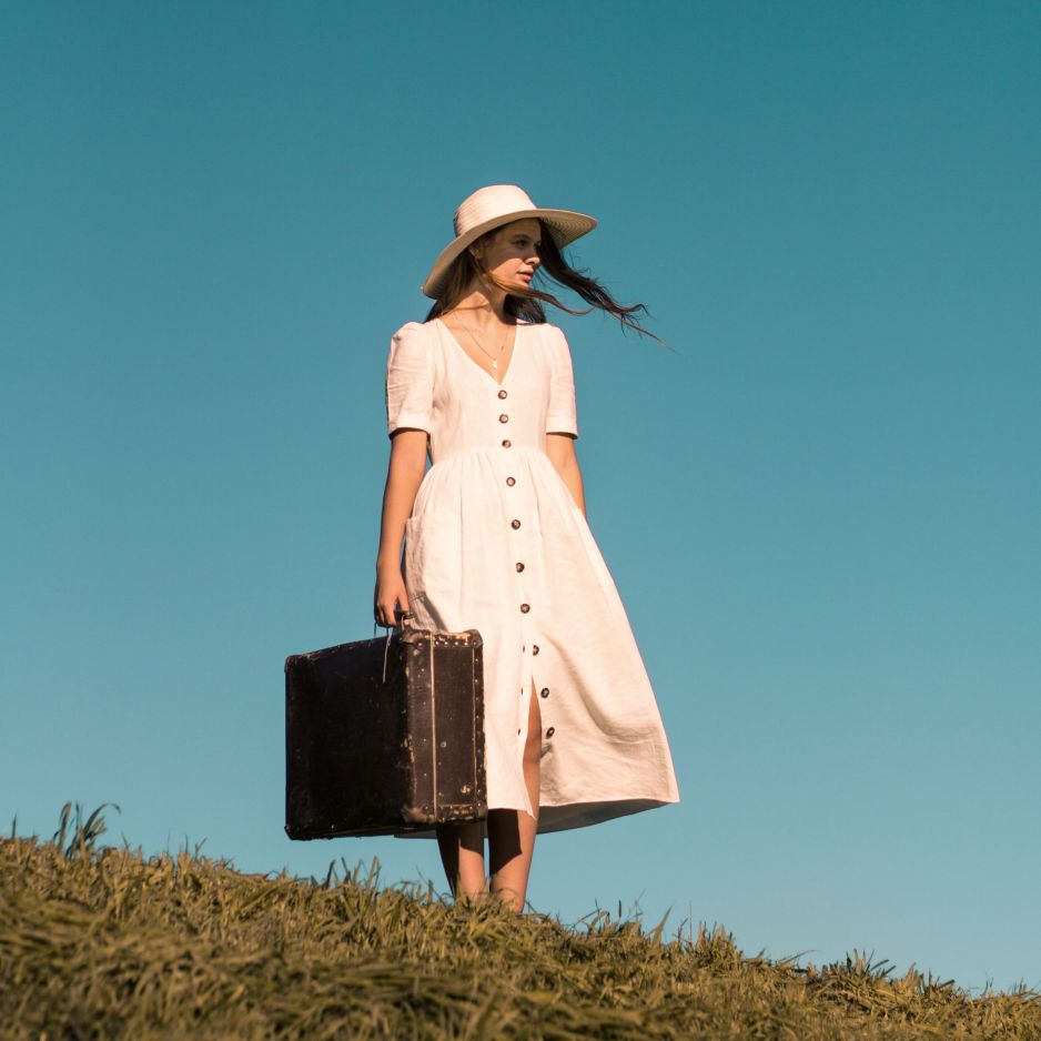 Eine Frau im Sommerkleid und mit Hut steht mit einem Koffer in der Hand auf einer Wiese